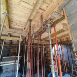 Wnętrze konstrukcji szalunkowej stropu z drewnianym deskowaniem, podpartej stalowymi stemplami o różnej kolorystyce, widoczne bloczki betonowe i osoba siedząca w tle.