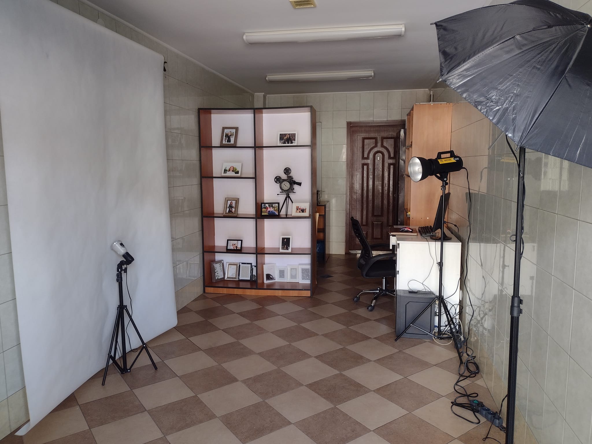 Studio fotograficzne z białym tłem, lampą błyskową z parasolem, regałem z ramkami i rekwizytami, oraz szachownicą na podłodze.