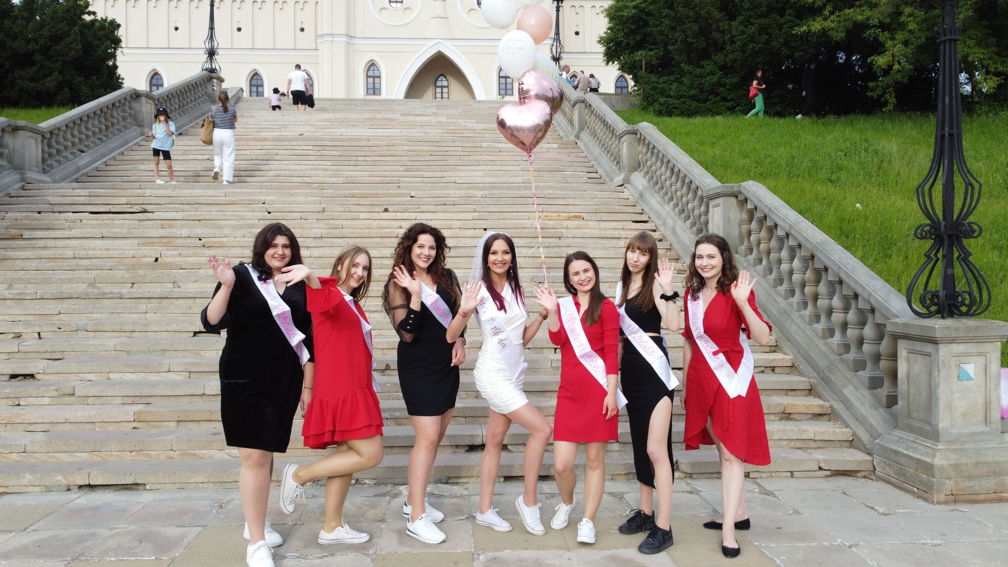 Grupa kobiet świętuje wieczór panieński na schodach z balustradą, trzymając balony i nosząc szarfy z napisem 'Wieczór Panieński'