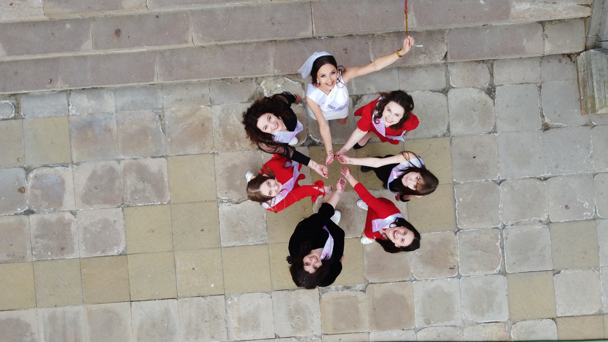 Zdjęcie z góry przedstawia grupę sześciu uśmiechniętych kobiet na zewnątrz, prawdopodobnie podczas wieczoru panieńskiego, gdzie panna młoda w białej sukience i welonie trzyma w ręku balon,...