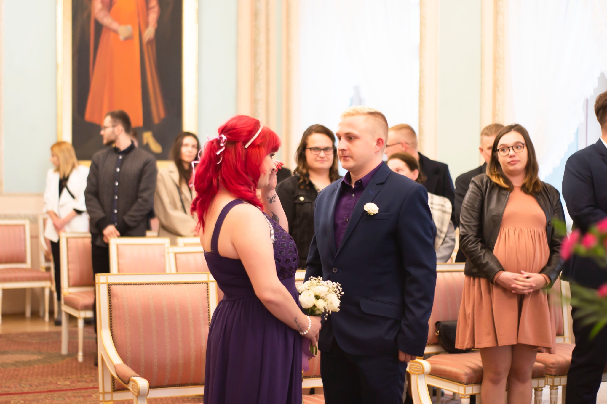 Panna młoda z czerwonymi włosami ociera łzę wzruszenia podczas ceremonii ślubnej, w tle goście i obraz.