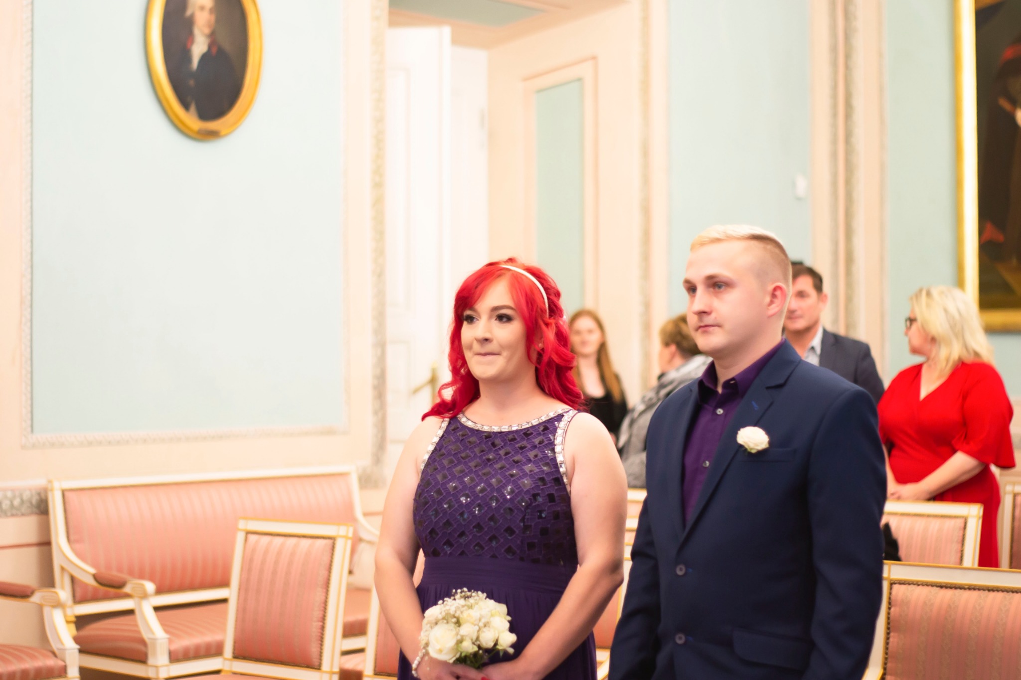 Para młoda w eleganckich strojach stoi w sali ślubów, panna młoda z rudymi włosami trzyma bukiet białych róż, w tle goście i portret na ścianie.