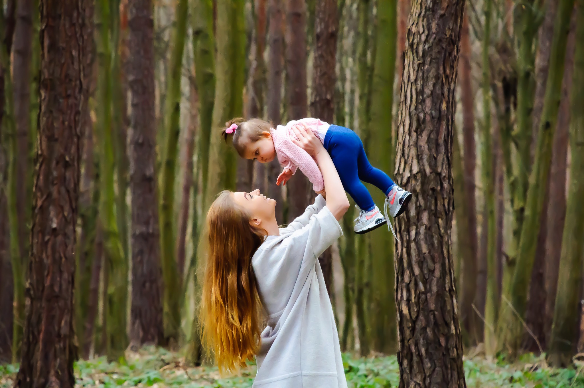 Matka podnosi córeczkę do góry w lesie sosnowym, patrząc na nią z uśmiechem, w tle widoczne drzewa i ściółka leśna.