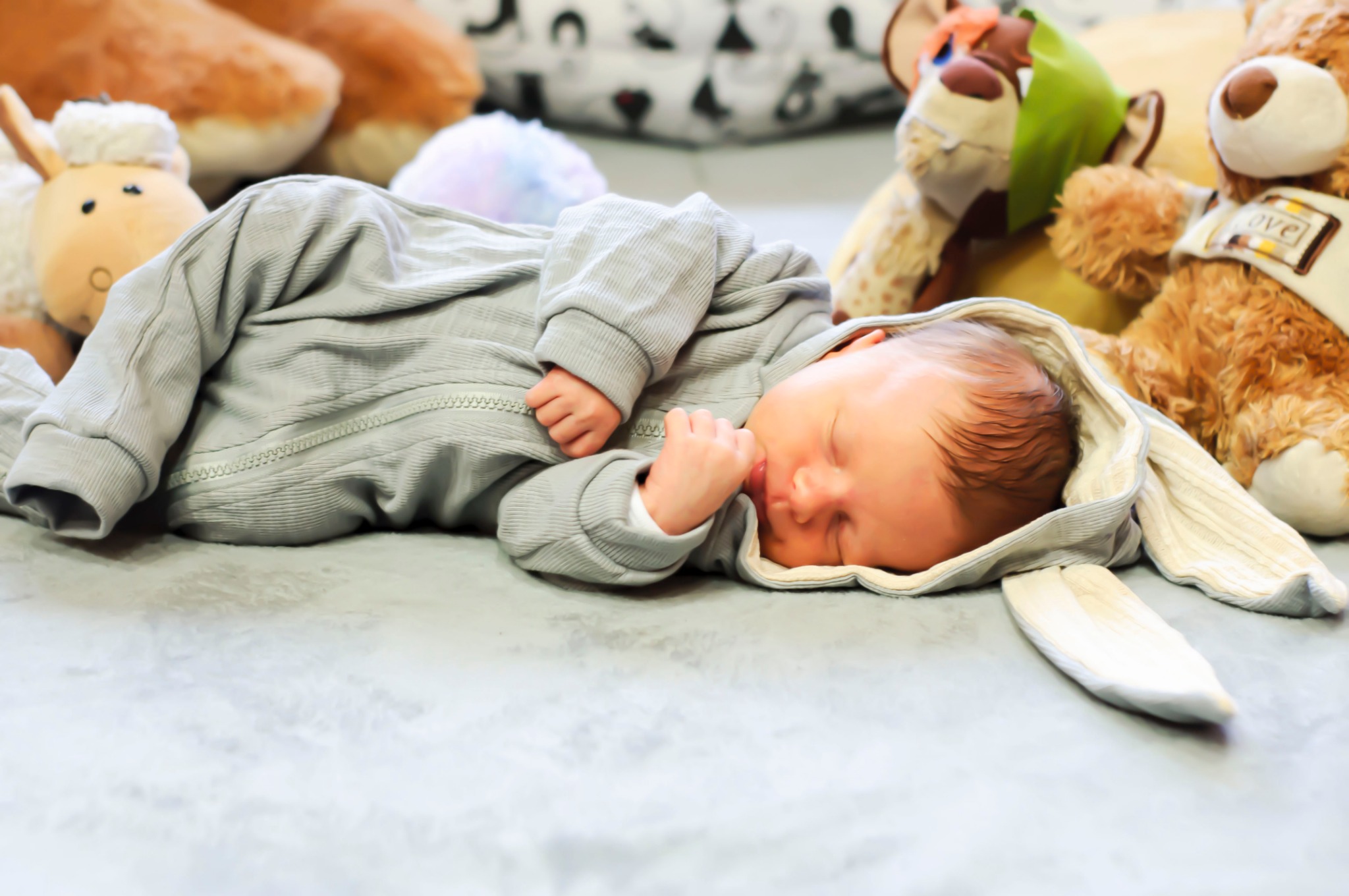 Śpiący noworodek w szarym kombinezonie z kapturem w kształcie króliczych uszu, otoczony pluszowymi zabawkami na miękkim, jasnym tle.