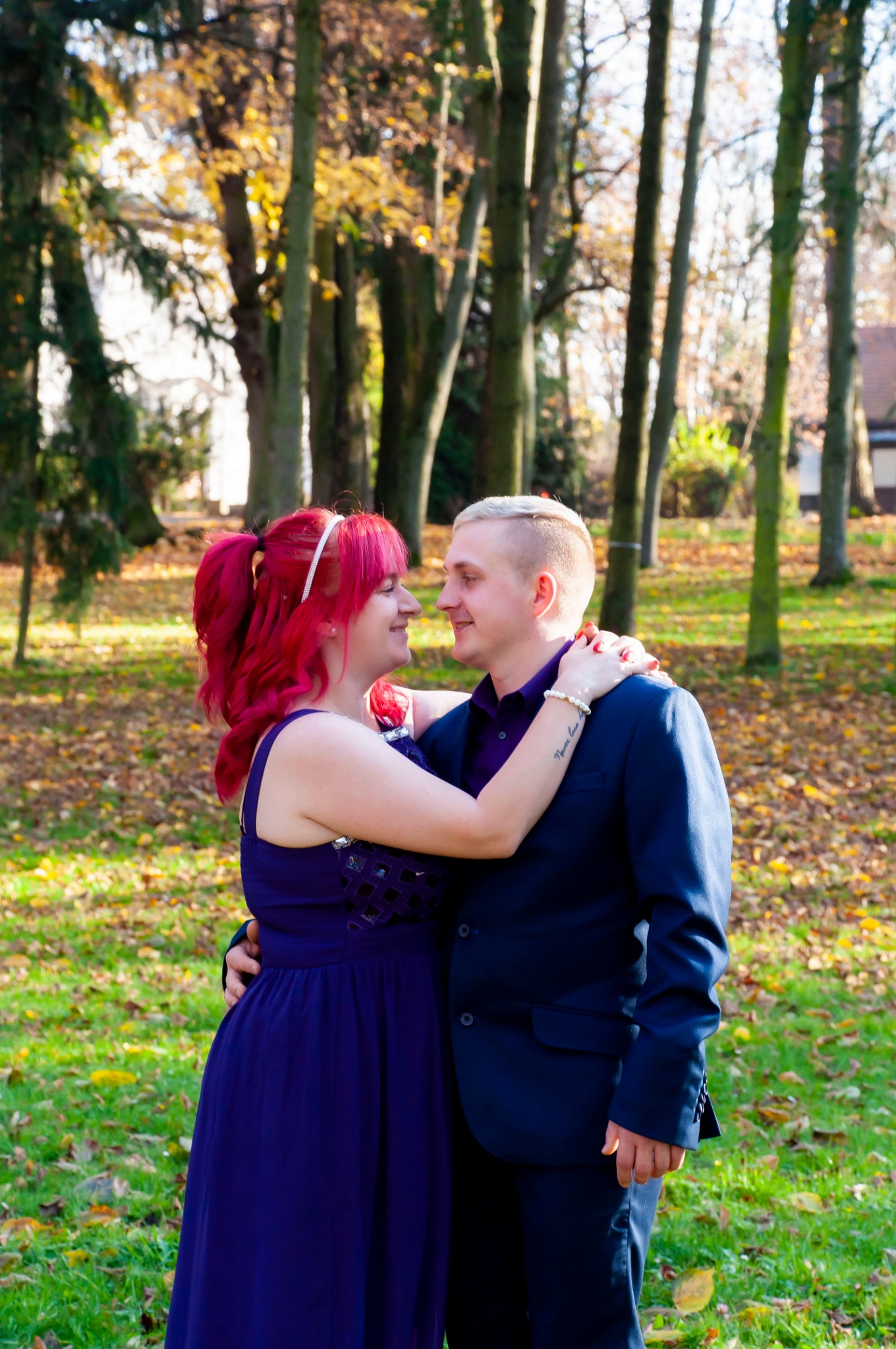 Zakochana para w objęciach na tle jesiennego parku, ona w długiej fioletowej sukni z cekinami, on w granatowym garniturze i fioletowej koszuli, oboje uśmiechnięci.