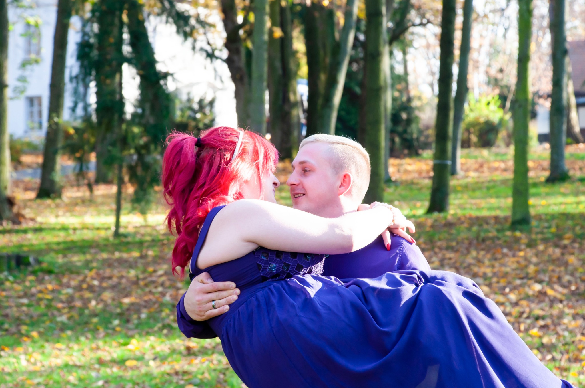 Uśmiechnięty mężczyzna trzyma na rękach kobietę z różowymi włosami w granatowej sukience na tle parku jesienią.