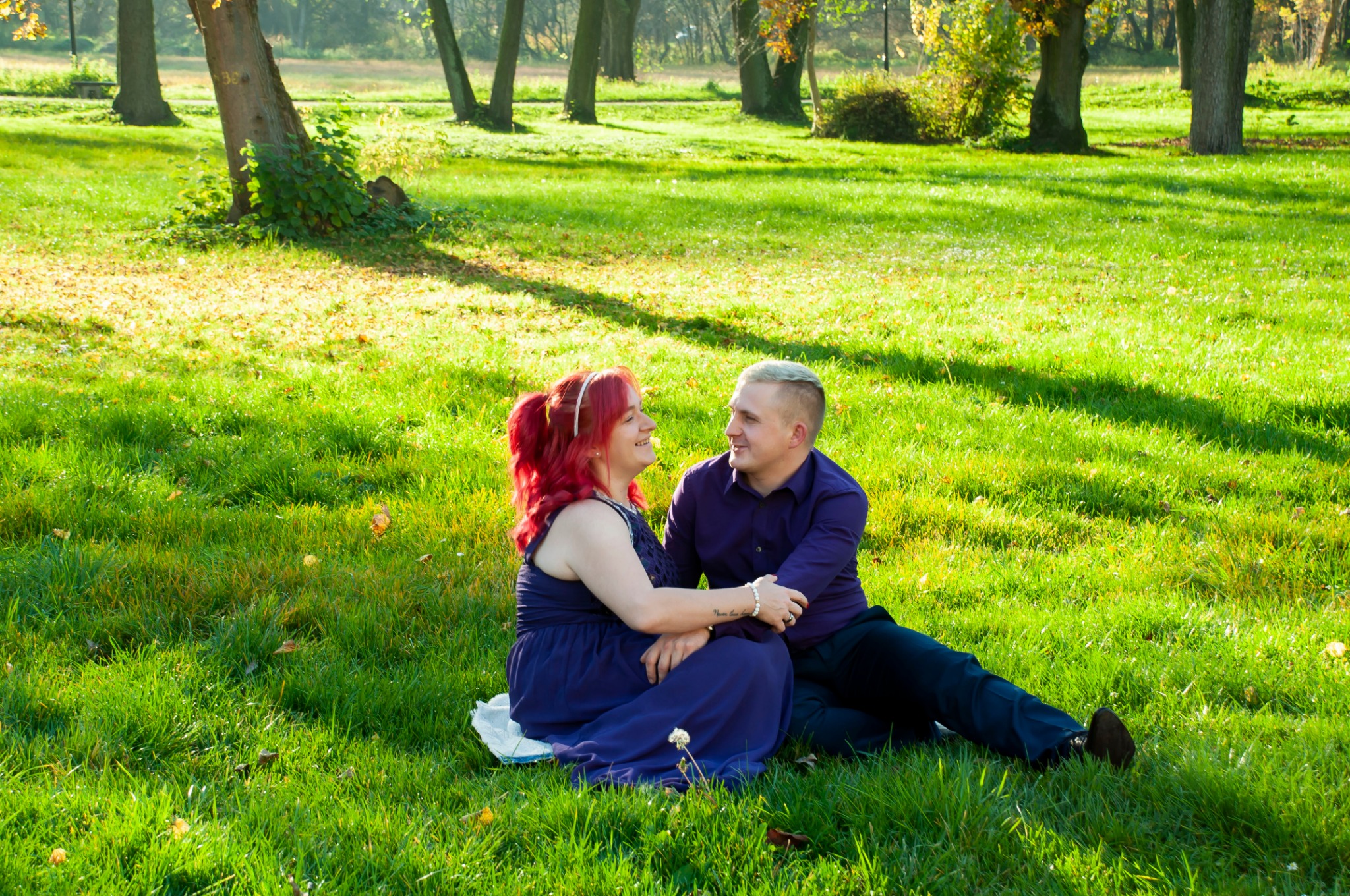 Uśmiechnięta para siedzi na trawie w parku, kobieta z czerwonymi włosami i mężczyzna z blond fryzurą patrzą na siebie z czułością, otoczeni zielenią i drzewami w słoneczny dzień.
