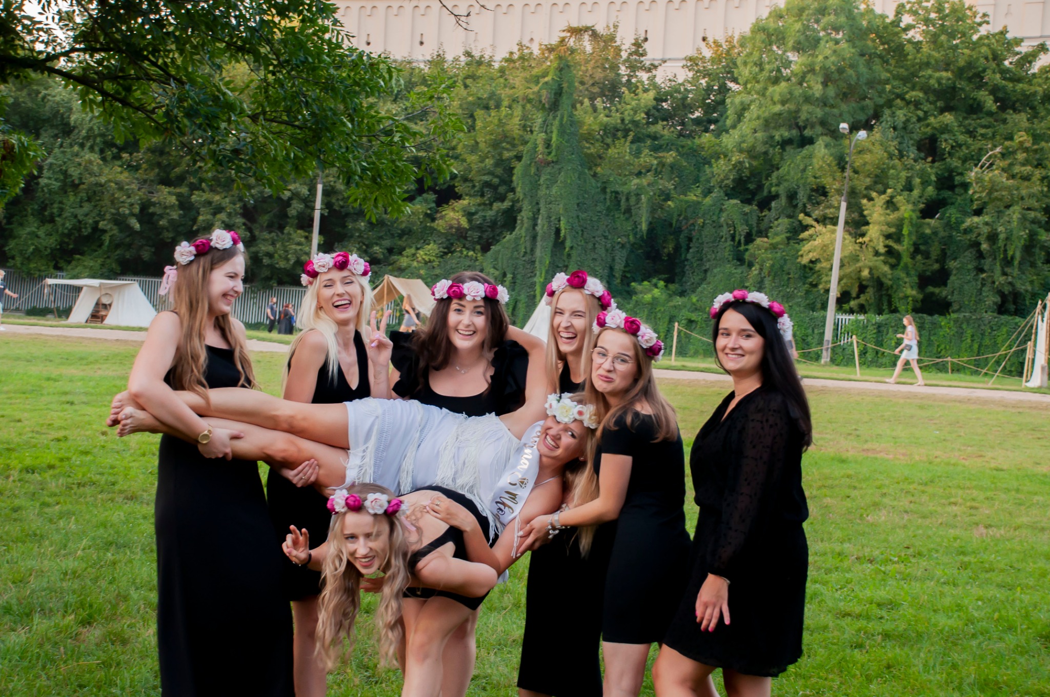 Grupa roześmianych kobiet w czarnych sukienkach i wiankach na głowach, trzymająca pannę młodą w białej sukience, na tle zieleni i namiotów.