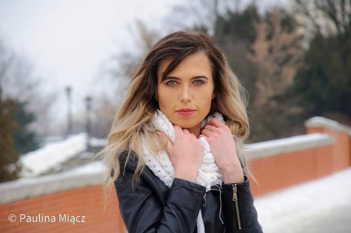 Portret kobiety w czarnej skórzanej kurtce i białym szaliku na tle zimowego krajobrazu z ceglanym murem.