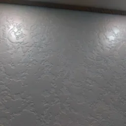 Szara ściana z dekoracyjną fakturą przypominającą nierówny tynk, oświetlona z góry, z widoczną liną dekoracyjną przy suficie.