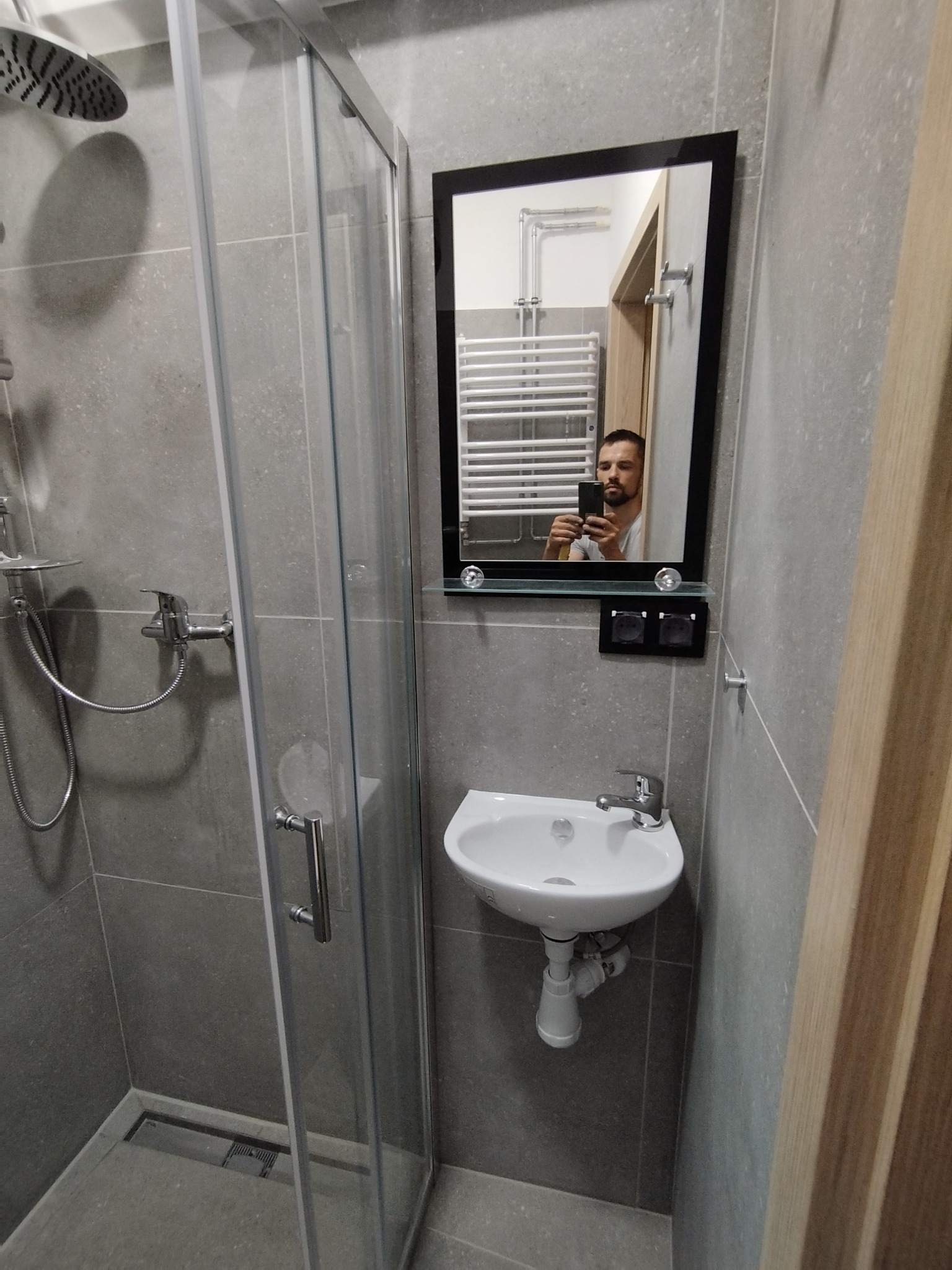 Nowoczesna łazienka w odcieniach szarości z kabiną prysznicową typu walk-in i małą umywalką; widoczne odbicie fotografa w lustrze.