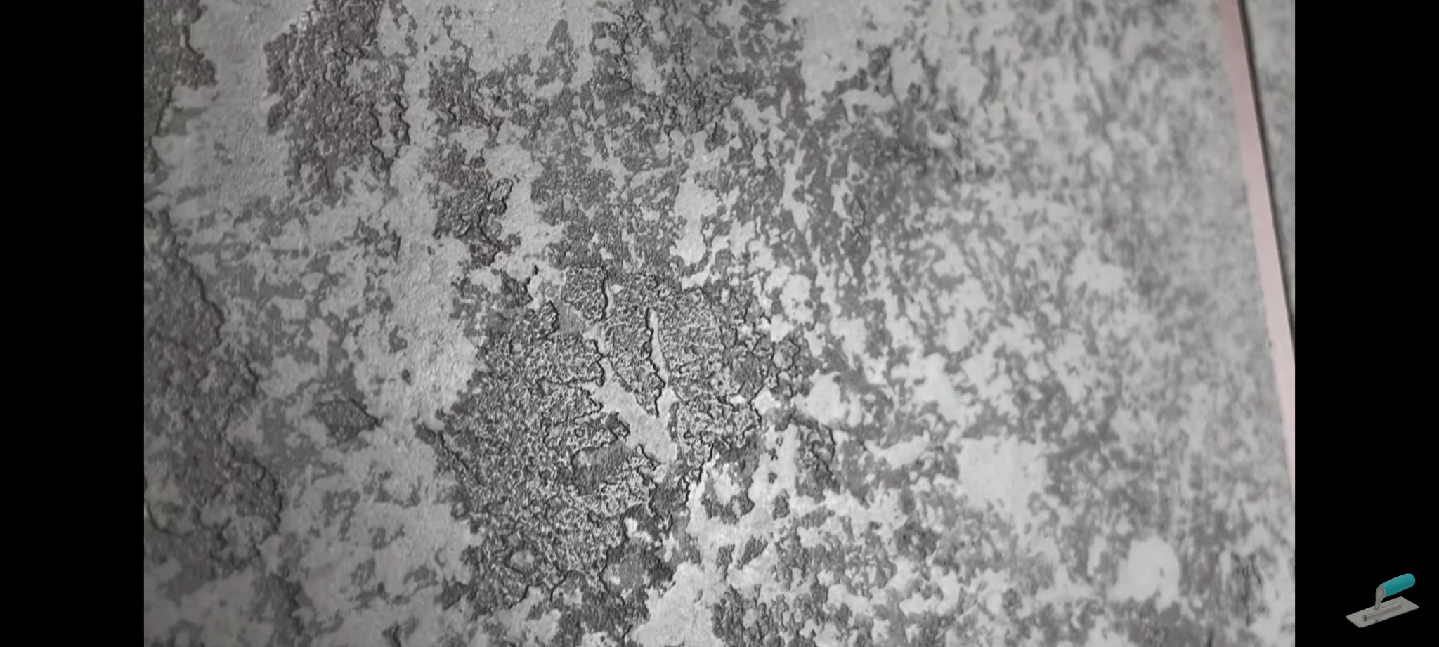 Szara ściana z dekoracyjną, nieregularną fakturą przypominającą płatki farby, widoczny fragment listwy wykończeniowej.