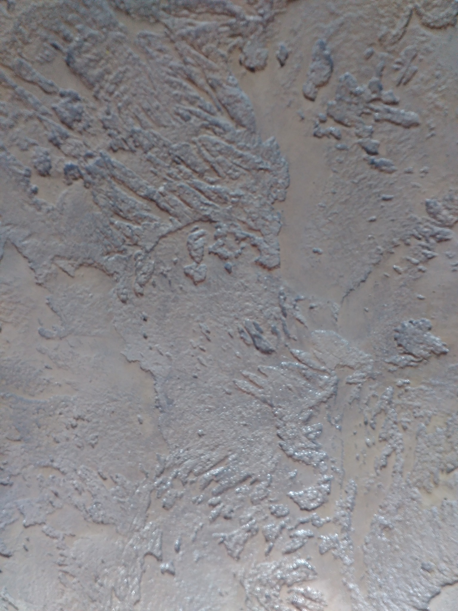 Szara, teksturowana ściana z wyraźnymi, nieregularnymi wzorami przypominającymi pociągnięcia pędzla, lekko połyskująca w świetle.