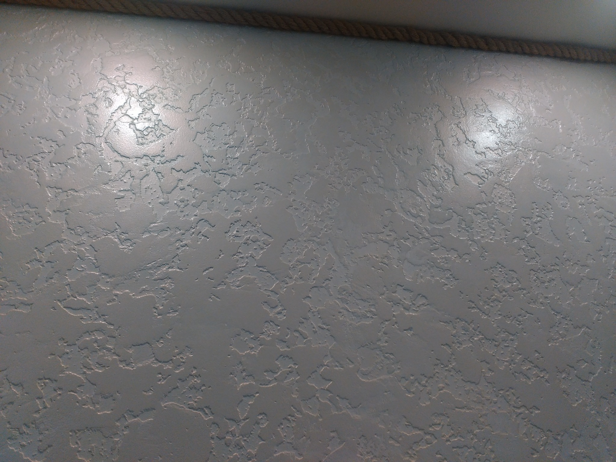 Szara ściana z dekoracyjną fakturą przypominającą nierówny tynk, oświetlona z góry, z widoczną liną dekoracyjną przy suficie.