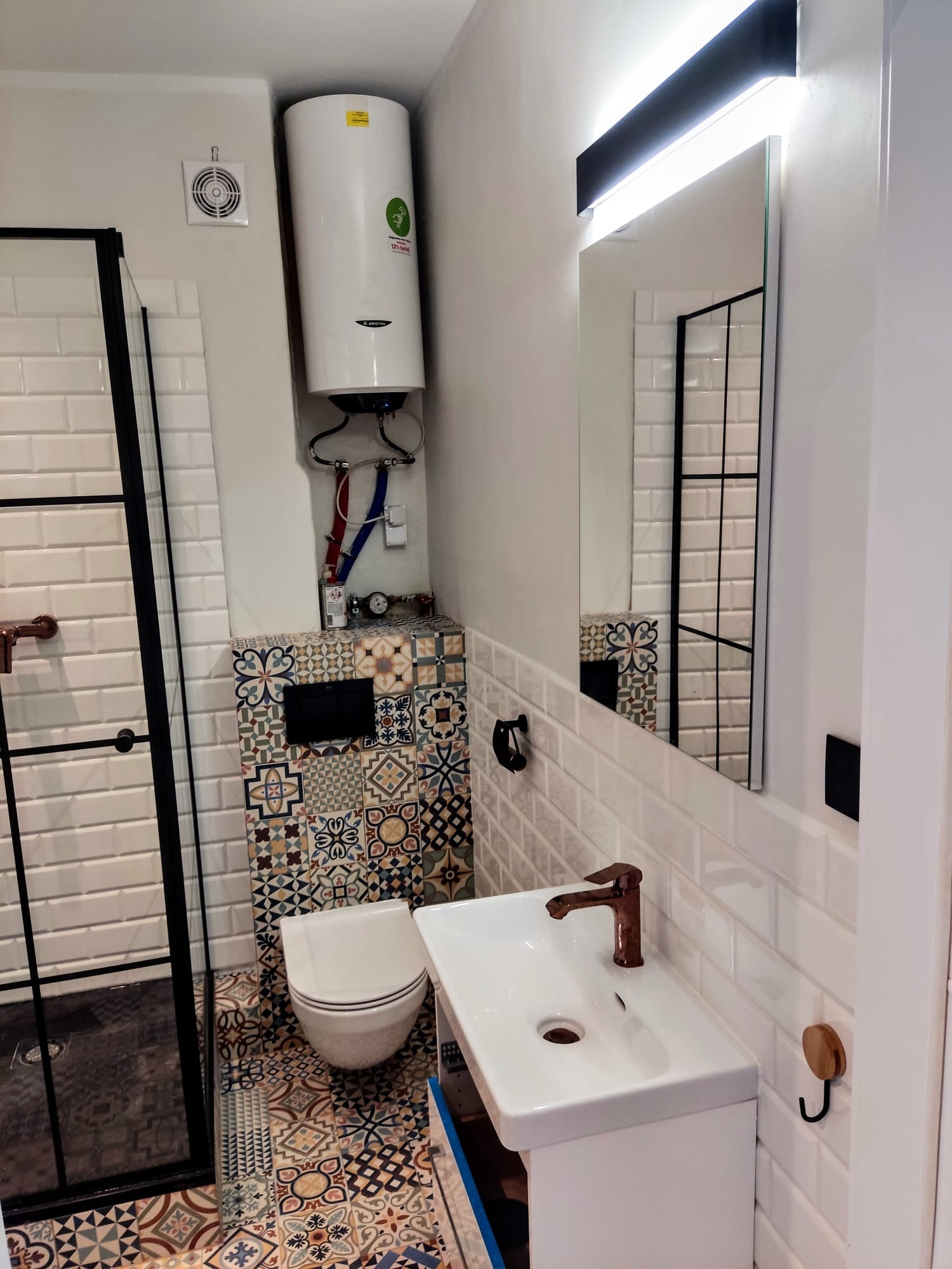 Nowoczesna łazienka z kabiną prysznicową z czarnymi ramami, wiszącą toaletą, umywalką z brązową armaturą, wzorzystymi płytkami na podłodze i ścianie za toaletą oraz białymi płytkami imitującymi...