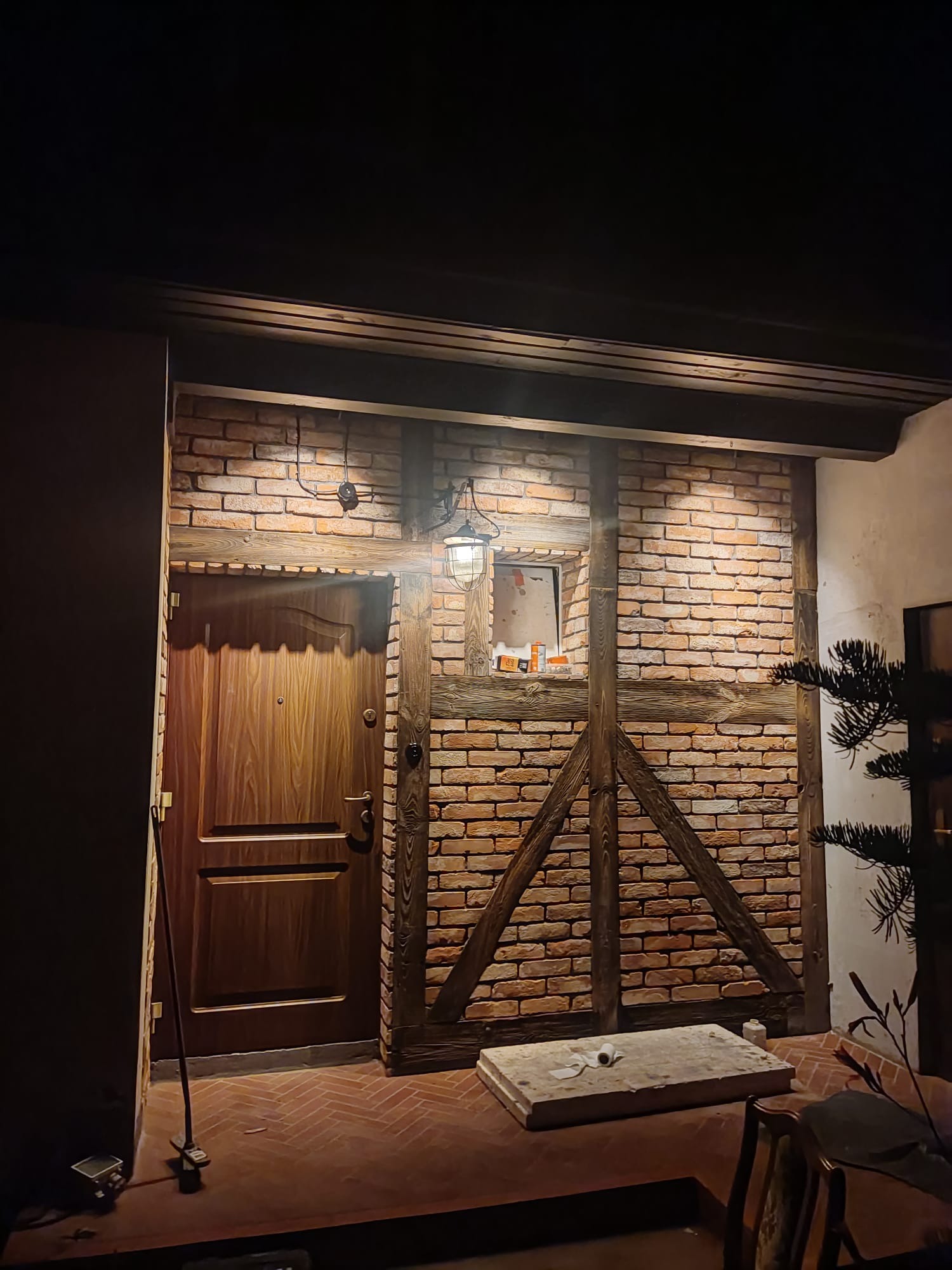 Wejście do budynku w stylu rustykalnym z drewnianymi belkami i ceglaną ścianą, oświetlone wiszącą lampą w stylu retro, widoczna drewniana brama i fragment podłogi z cegły