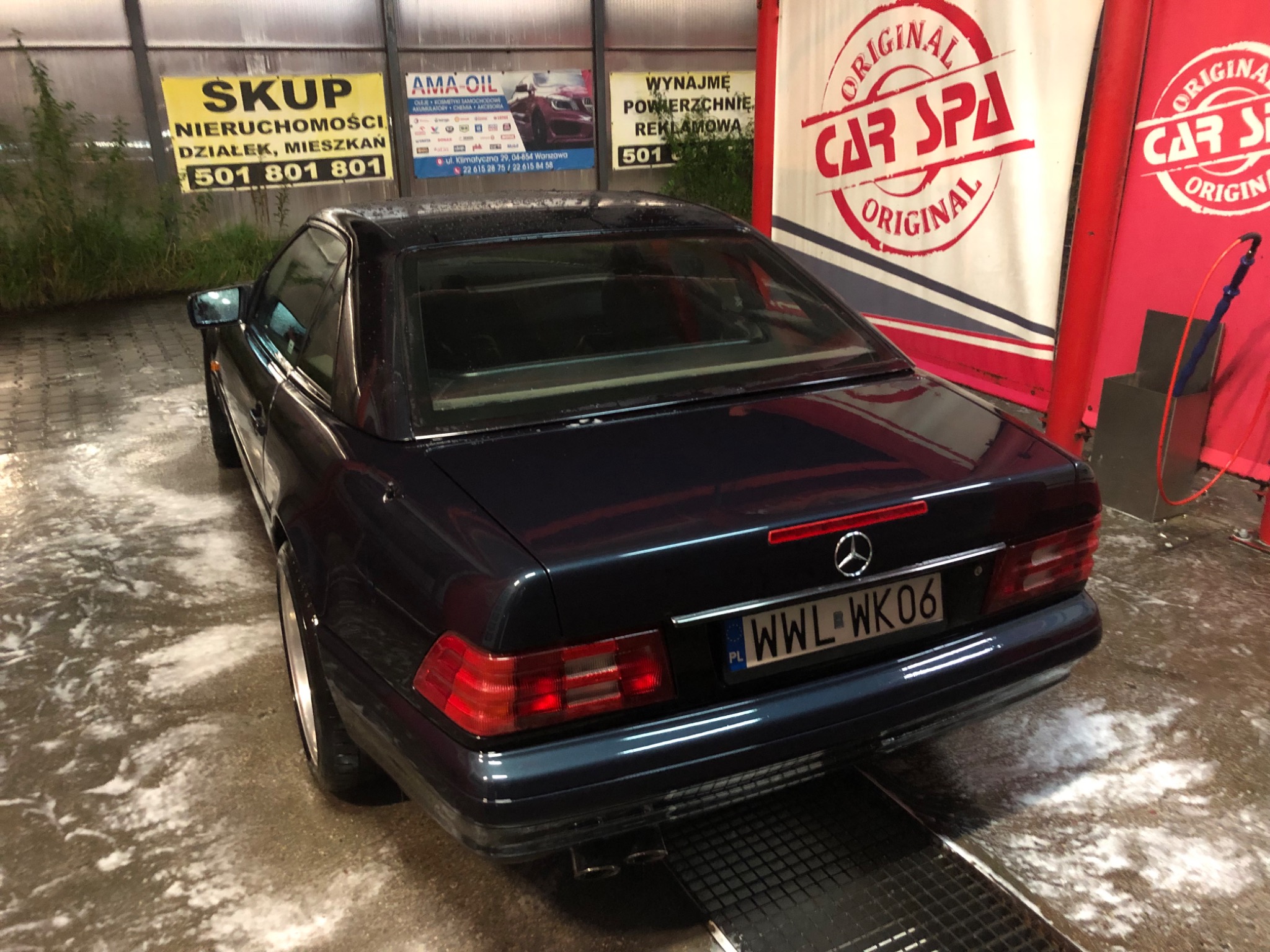 Czarny Mercedes SL (R129) po myciu w myjni samochodowej, widok z tyłu, mokra karoseria i podłoże, w tle reklamy i szyldy.