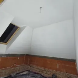 Pokój na poddaszu w trakcie prac wykończeniowych: ściany i sufit po nałożeniu gładzi, widoczne cegły na dole ścian zabezpieczone folią, okno dachowe