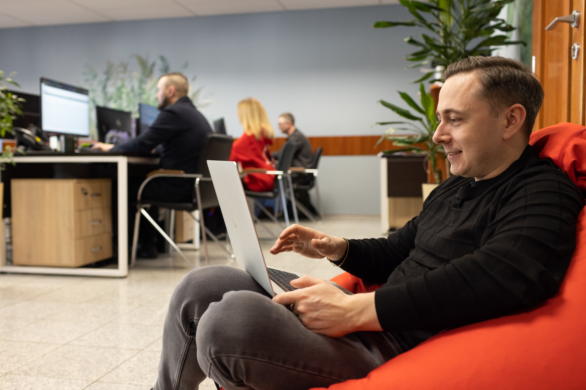 Uśmiechnięty mężczyzna w czarnym swetrze siedzi na czerwonym pufie, pracując na srebrnym laptopie w biurze z innymi osobami pracującymi przy biurkach w tle.