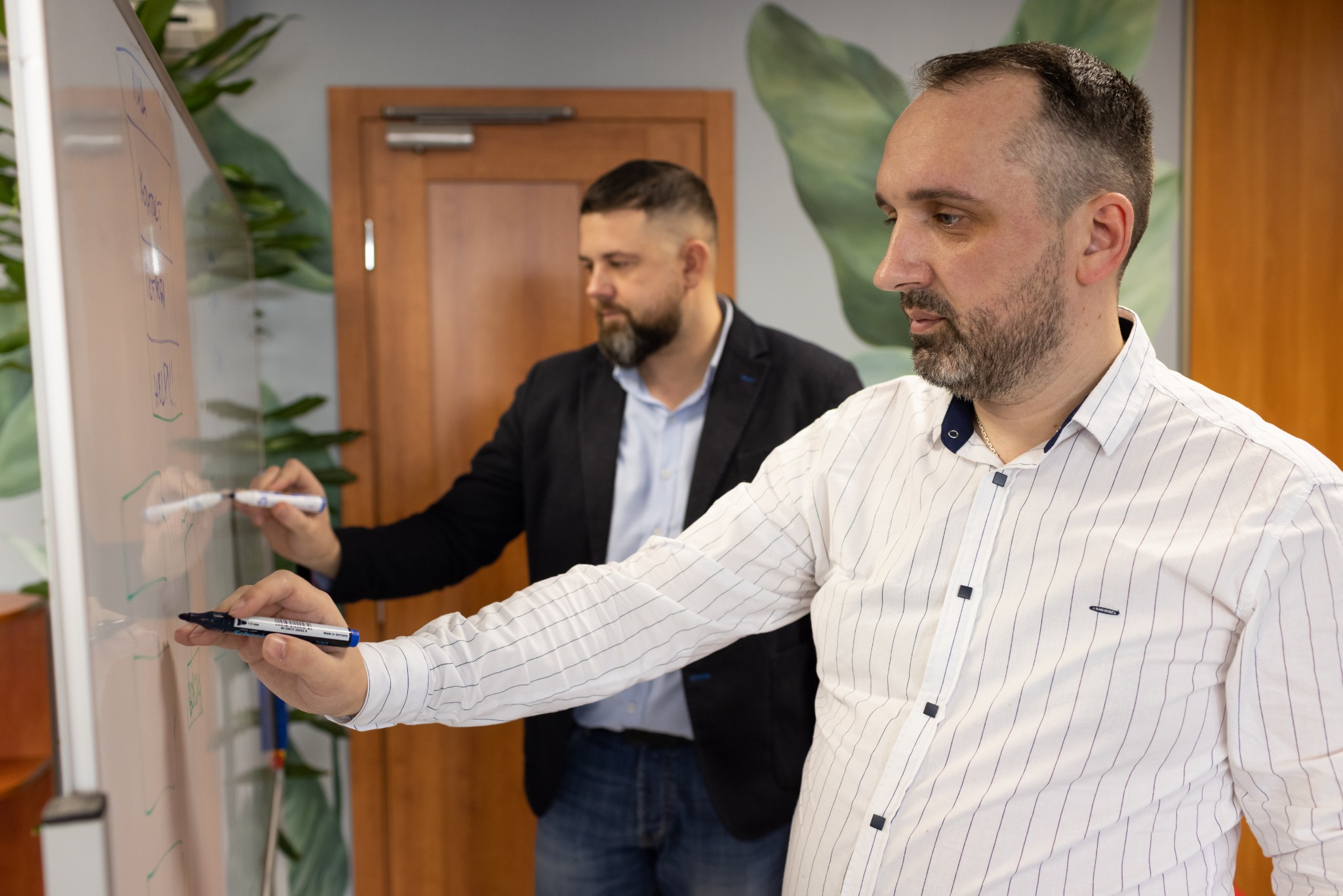 Dwóch mężczyzn pisze markerami na białej tablicy, prawdopodobnie omawiając strukturę lub schemat działania, widoczny fragment napisu 'HELP!!'.