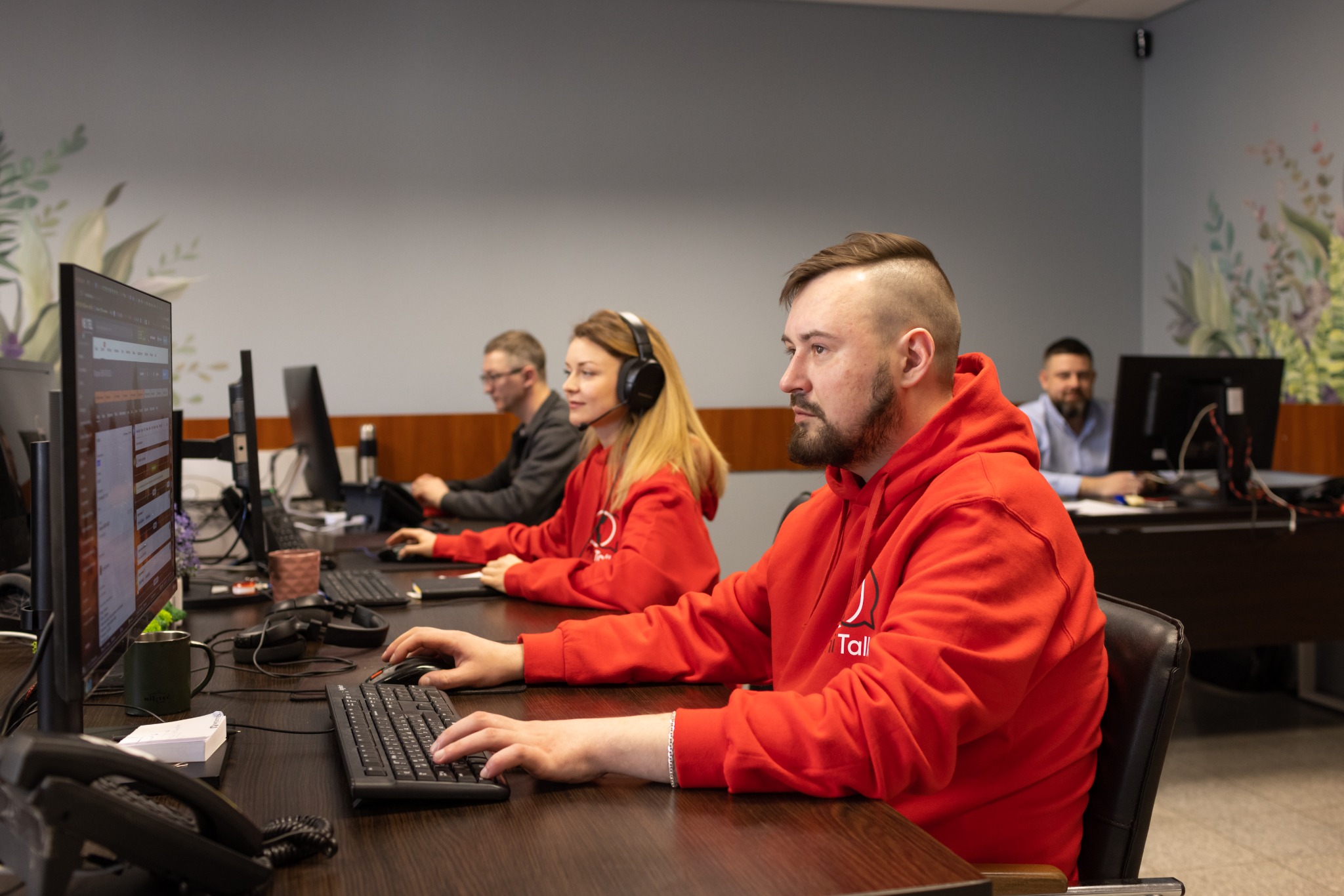 Zespół pracowników w biurze, korzystający z komputerów, prawdopodobnie pracujący nad systemem telefoni internetowej, mężczyzna w czerwonej bluzie z logo firmy na pierwszym planie.