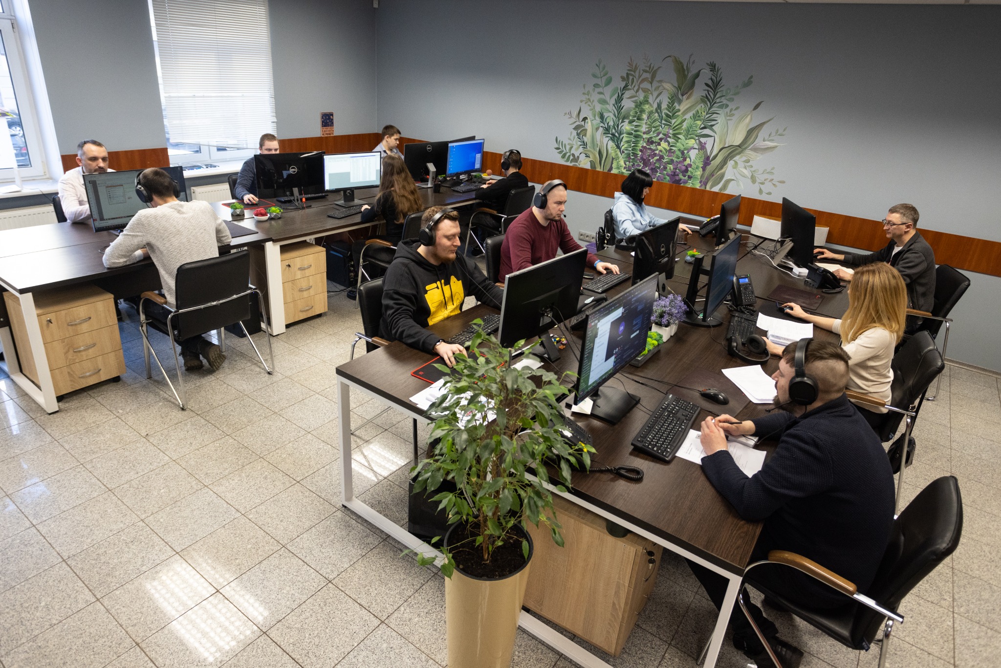 Biuro obsługi klienta: pracownicy w słuchawkach pracują przy komputerach, na ścianie widoczne dekoracje roślinne.