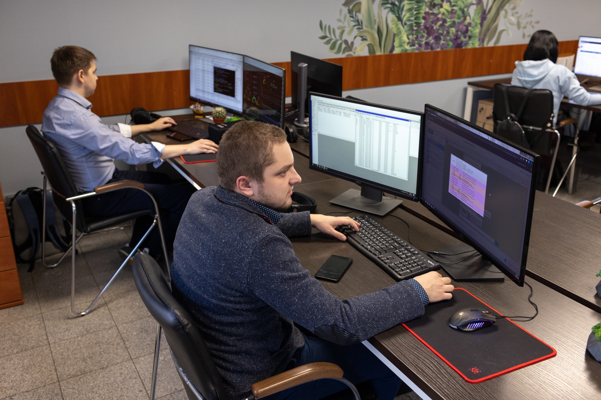 Programiści pracują przy biurkach z dwoma monitorami, analizując kod; widoczny fragment biura z dekoracyjną ścianą w roślinne motywy.