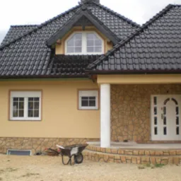 Żółta elewacja domu jednorodzinnego z kamienną podmurówką i wejściem, czarny dach z dachówki, taczka stoi przed budynkiem.