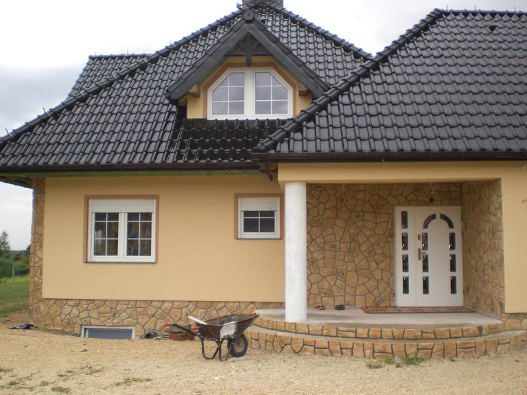 Żółta elewacja domu jednorodzinnego z kamienną podmurówką i wejściem, czarny dach z dachówki, taczka stoi przed budynkiem.