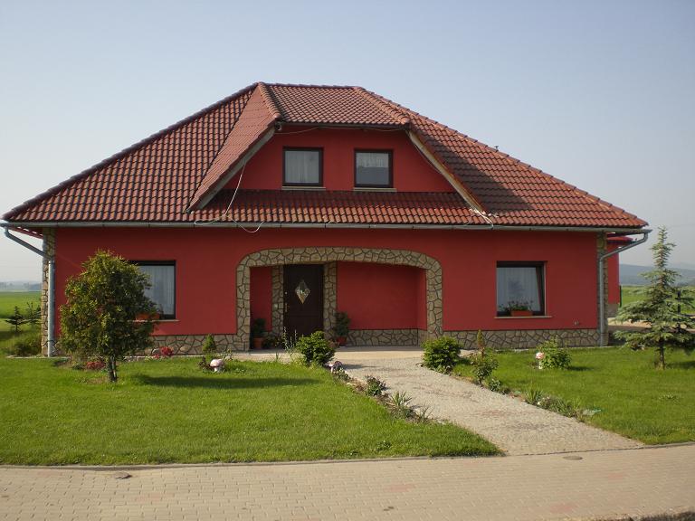 Czerwony dom jednorodzinny z brązową dachówką, kamiennym wykończeniem wokół wejścia i okien, oraz zadbanym trawnikiem.