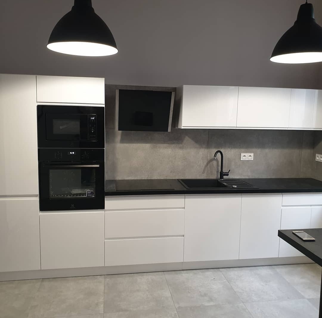 Nowoczesna kuchnia z białymi, błyszczącymi szafkami, czarnym blatem i sprzętem AGD w zabudowie, oświetlona wiszącymi lampami w kształcie kopuł.