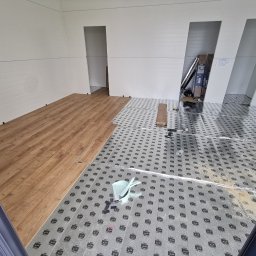 Jakość Zrobimy - Fragment pomieszczenia w trakcie montażu podłogi z paneli drewnopodobnych, widoczna podkładka pod panele z nadrukiem, białe panele ścienne i narzędzia