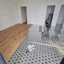 Fragment pomieszczenia w trakcie montażu podłogi z paneli drewnopodobnych, widoczna podkładka pod panele z nadrukiem, białe panele ścienne i narzędzia