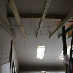 Jakość Zrobimy - Poddasze w trakcie prac wykończeniowych: zamontowane płyty gipsowo-kartonowe, widoczne drewniane belki konstrukcyjne, narzędzia na podłodze i stołek z wkrętarką przy oknie dachowym.