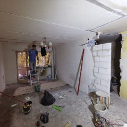 Jakość Zrobimy - Mężczyzna na drabinie w trakcie prac wykończeniowych w pomieszczeniu z odsłoniętą cegłą i przygotowaną ścianą, widoczne narzędzia i materiały budowlane.