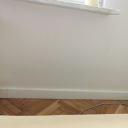 Jakość Zrobimy - Fragment pokoju po remoncie: parkiet w jodełkę, białe listwy przypodłogowe, grzejnik żeliwny i gładka, pomalowana na biało ściana.