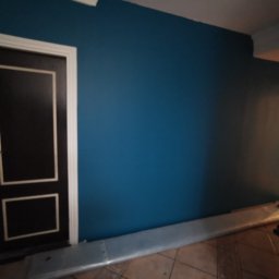 Jakość Zrobimy - Świeżo pomalowana, gładka ściana w odcieniu głębokiego turkusu z czarnymi drzwiami z białymi obramowaniami, widoczna listwa przypodłogowa w trakcie montażu i fragment brązowej podłogi.