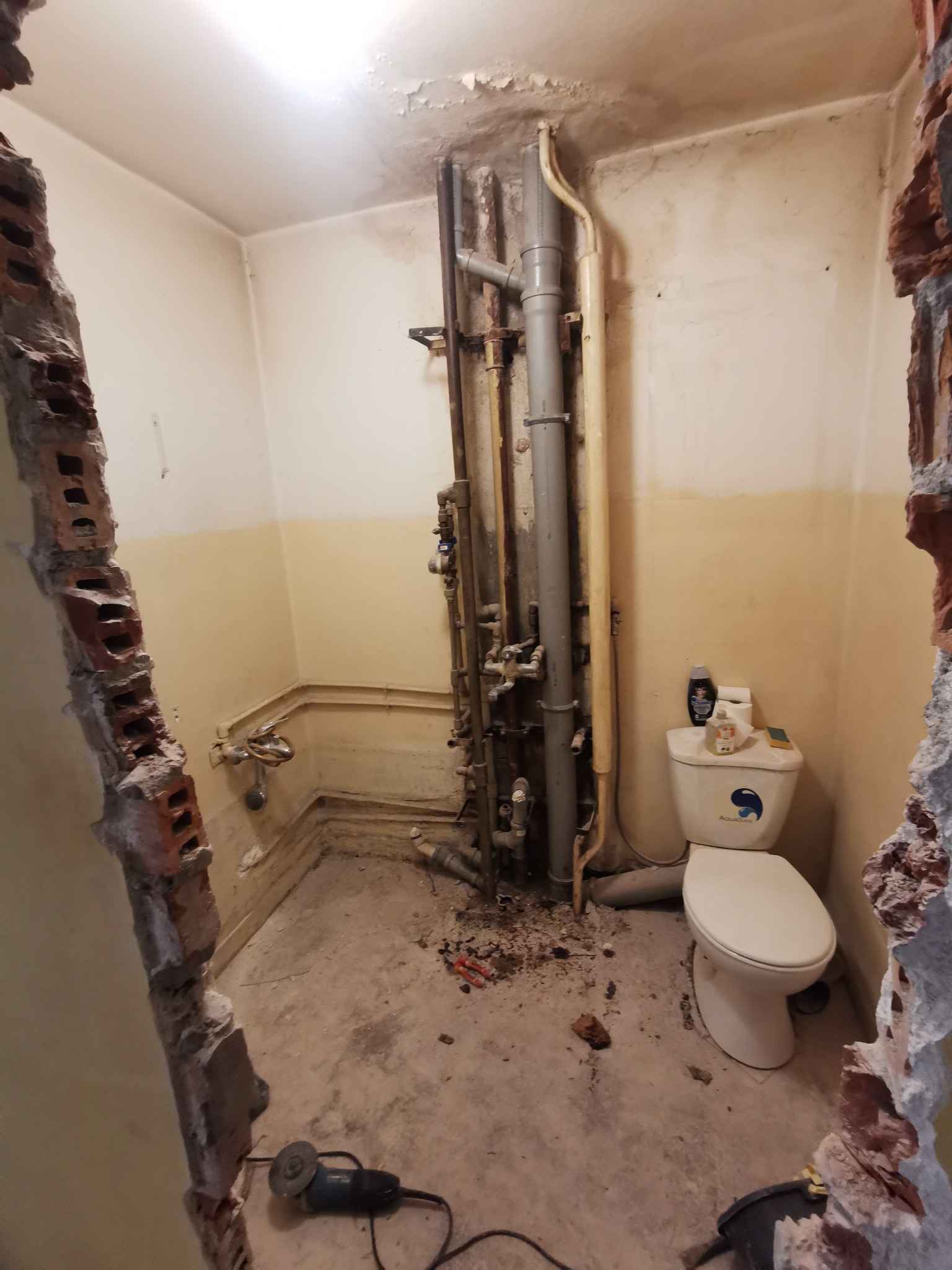 Łazienka w trakcie demontażu: widoczne odsłonięte rury, zniszczone ściany z fragmentami cegieł, stojąca toaleta oraz szlifierka kątowa na podłodze.