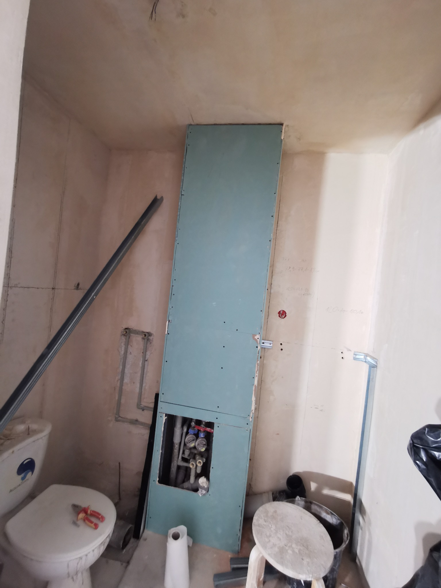 Niedokończona łazienka w trakcie remontu: widoczna instalacja wodno-kanalizacyjna za płytą kartonowo-gipsową, toaleta z narzędziami na desce, oraz surowe ściany z widocznymi notatkami i profilem...