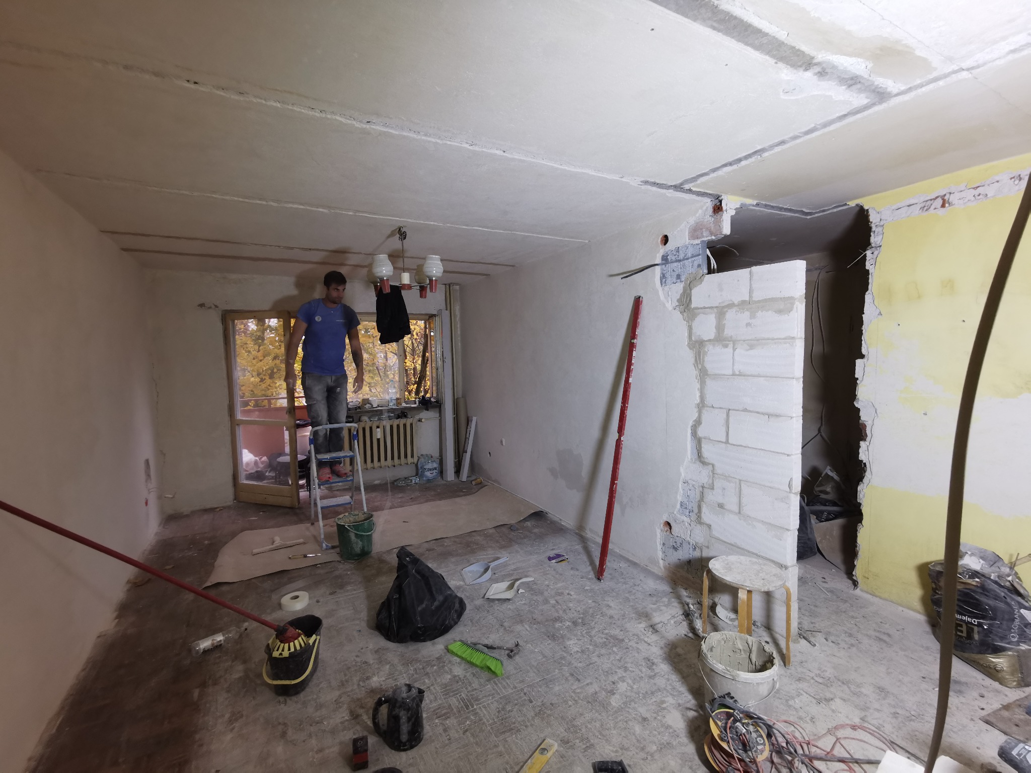 Mężczyzna na drabinie w trakcie prac wykończeniowych w pomieszczeniu z odsłoniętą cegłą i przygotowaną ścianą, widoczne narzędzia i materiały budowlane.