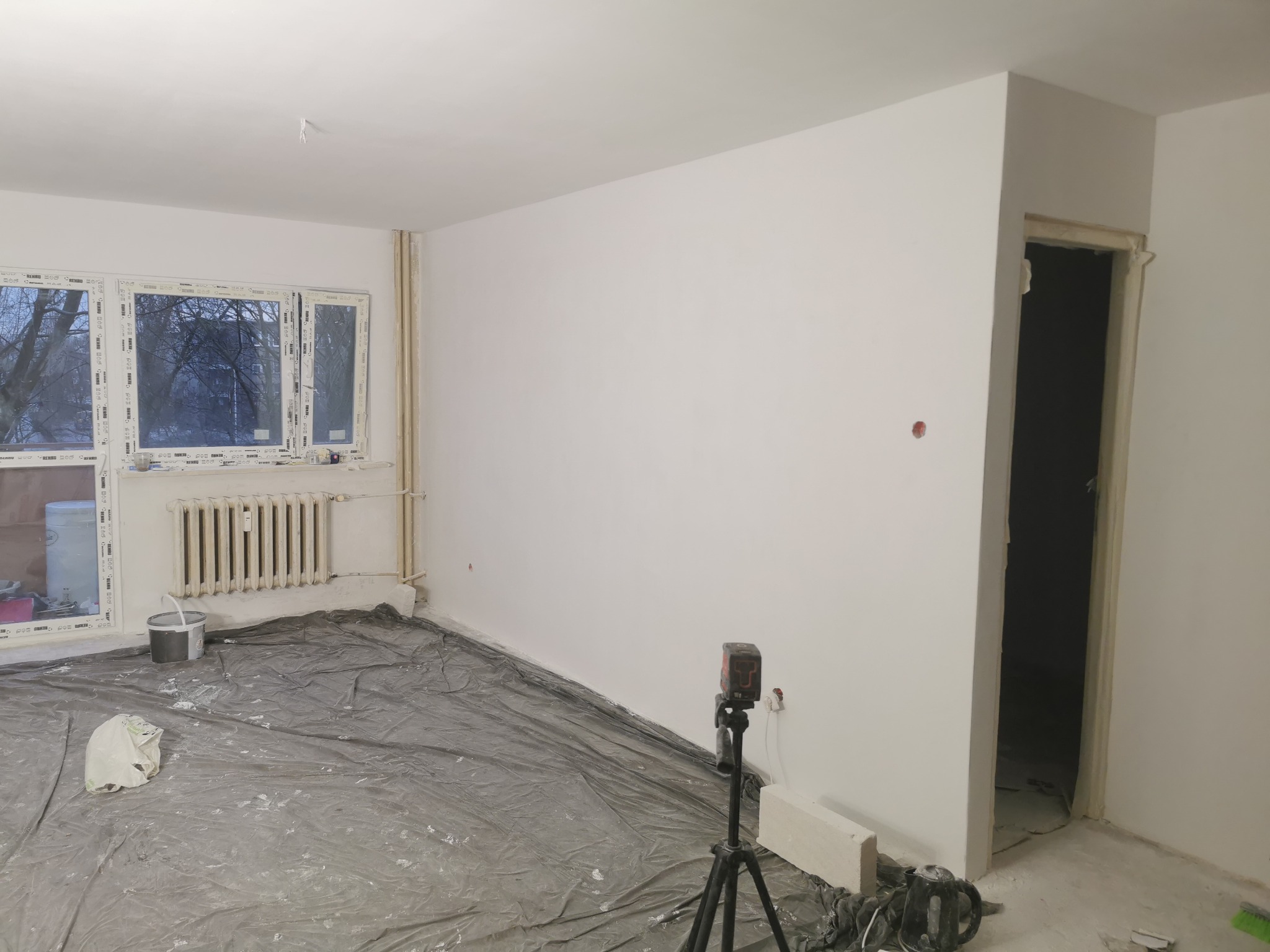 Pokój po świeżym malowaniu ścian na biało, z zabezpieczoną podłogą folią malarską, widoczny laser krzyżowy na statywie oraz grzejnik pod oknem.