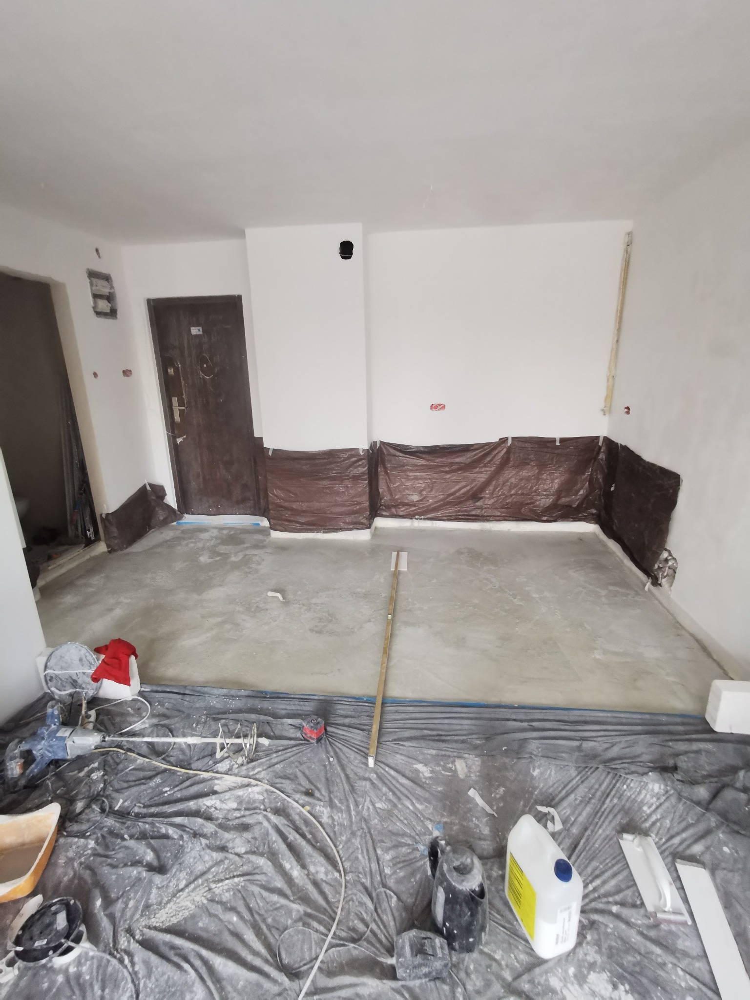 Przygotowanie do wylewki podłogowej w pomieszczeniu: zabezpieczone ściany folią, rozłożona folia ochronna, widoczny mikser elektryczny i pojemniki z zaprawą.