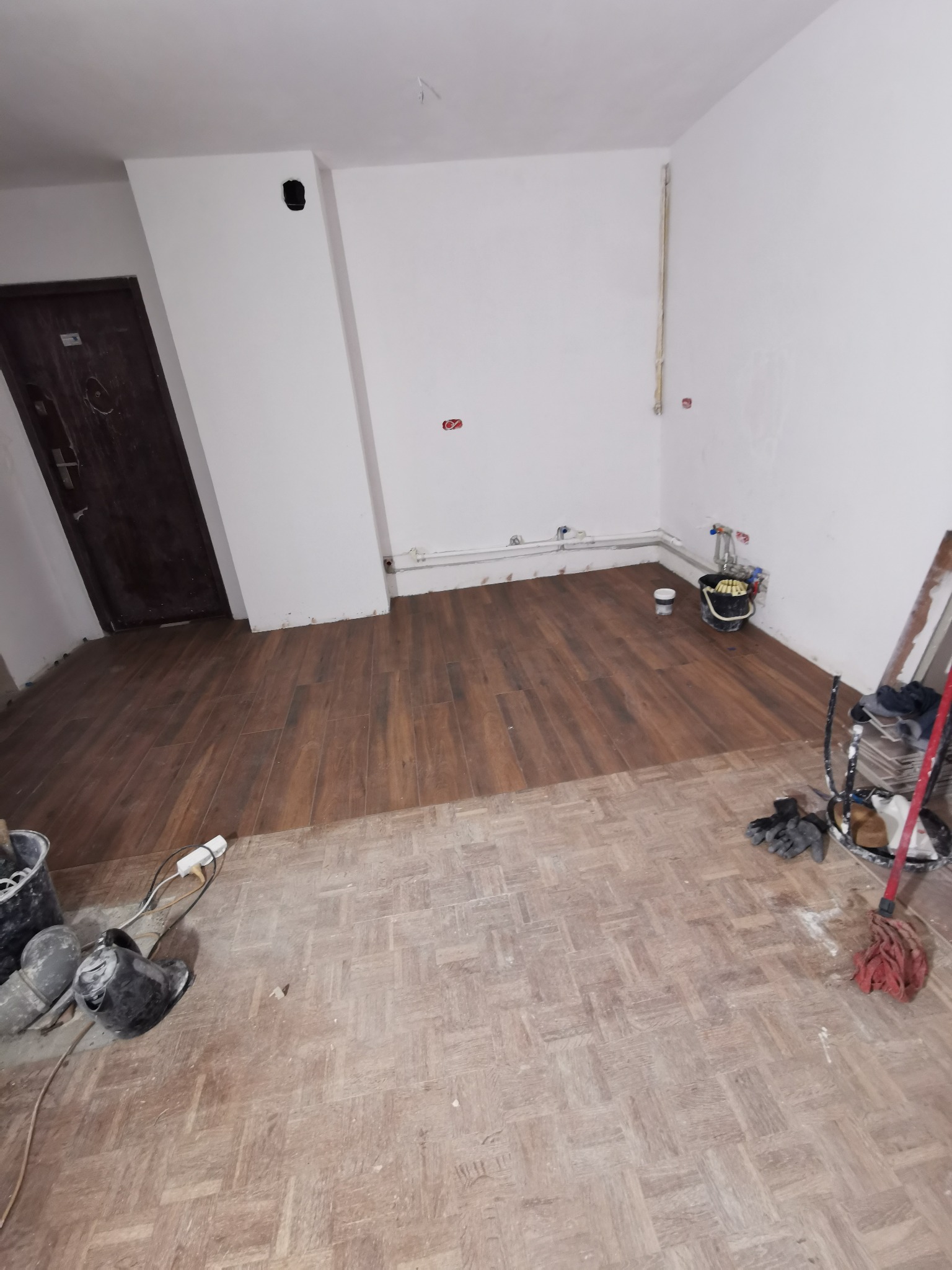 Fragment pokoju z częściowo ułożonymi panelami podłogowymi imitującymi drewno, widoczne stare panele, narzędzia i akcesoria glazurnicze.