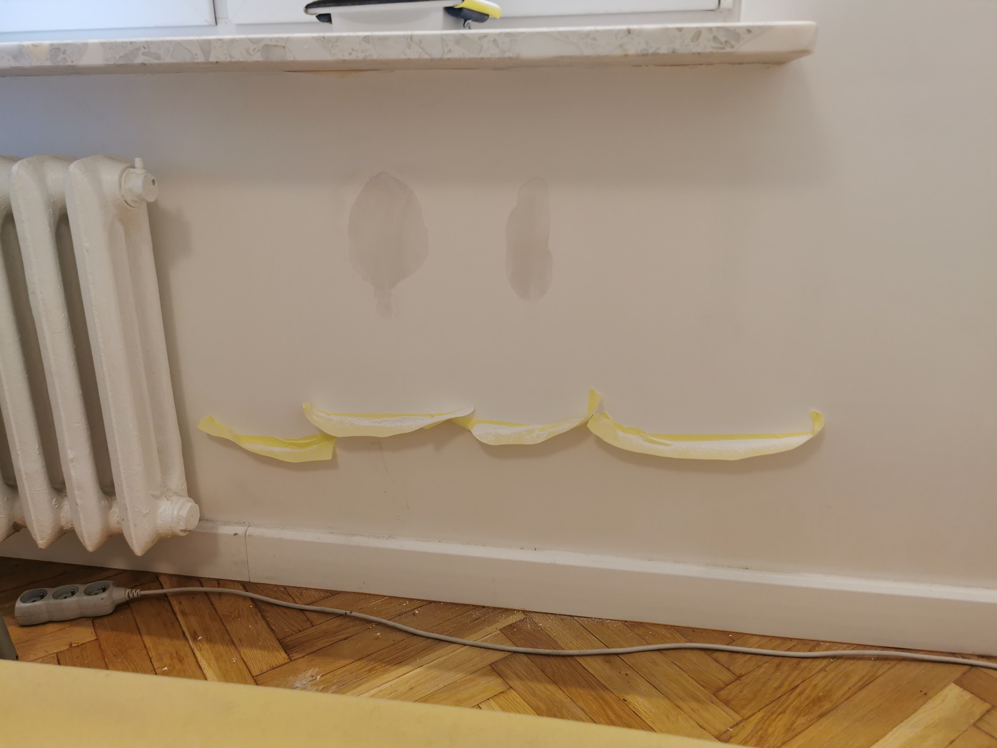 Ściana z widocznymi plamami po zalaniu, zabezpieczona taśmą malarską przed dalszymi pracami, z grzejnikiem i przedłużaczem przy podłodze z parkietu.
