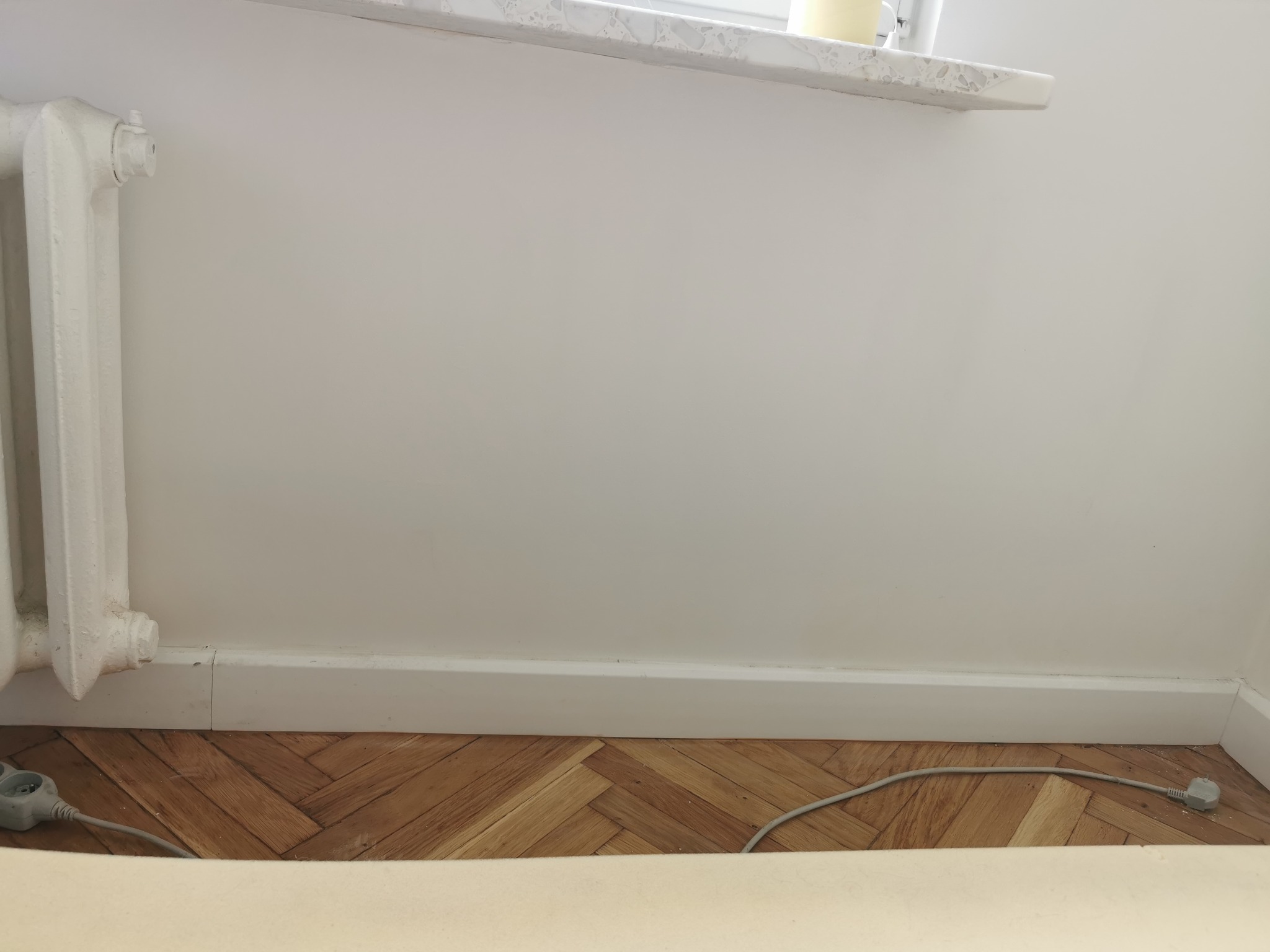Fragment pokoju po remoncie: parkiet w jodełkę, białe listwy przypodłogowe, grzejnik żeliwny i gładka, pomalowana na biało ściana.
