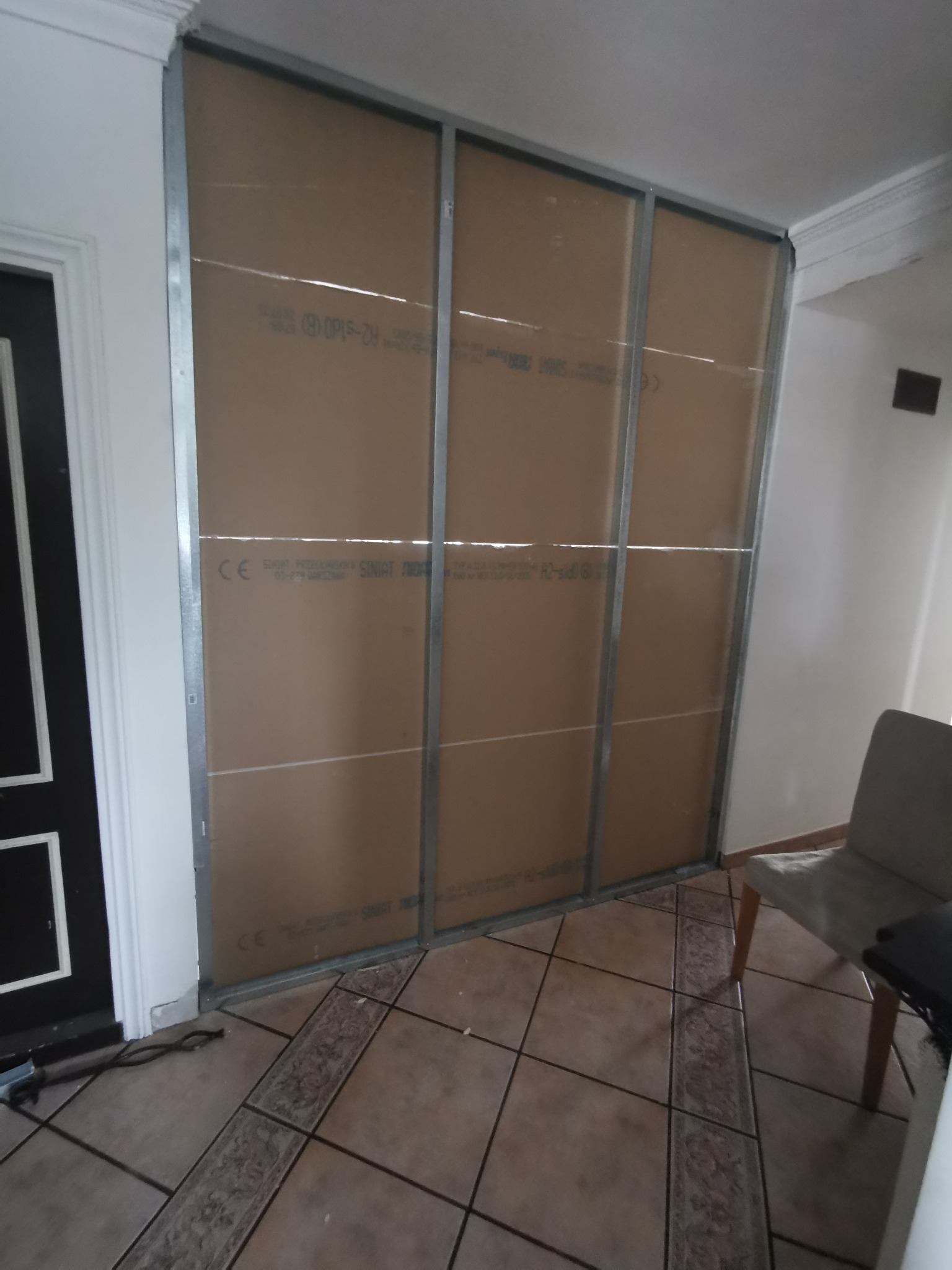 Konstrukcja ścianki działowej z profili stalowych i płyt kartonowo-gipsowych w trakcie montażu, widoczny fragment podłogi z płytek ceramicznych i krzesło w tle.