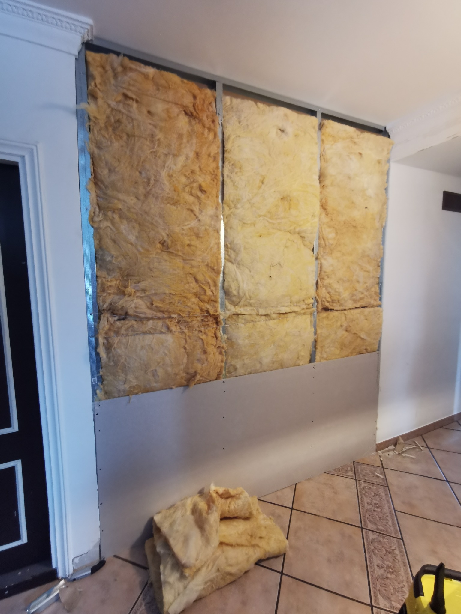 Montaż izolacji z wełny mineralnej w metalowym stelażu, częściowo zakrytej płytą gipsowo-kartonową, widoczny fragment podłogi z terakoty i narzędzie pracy.