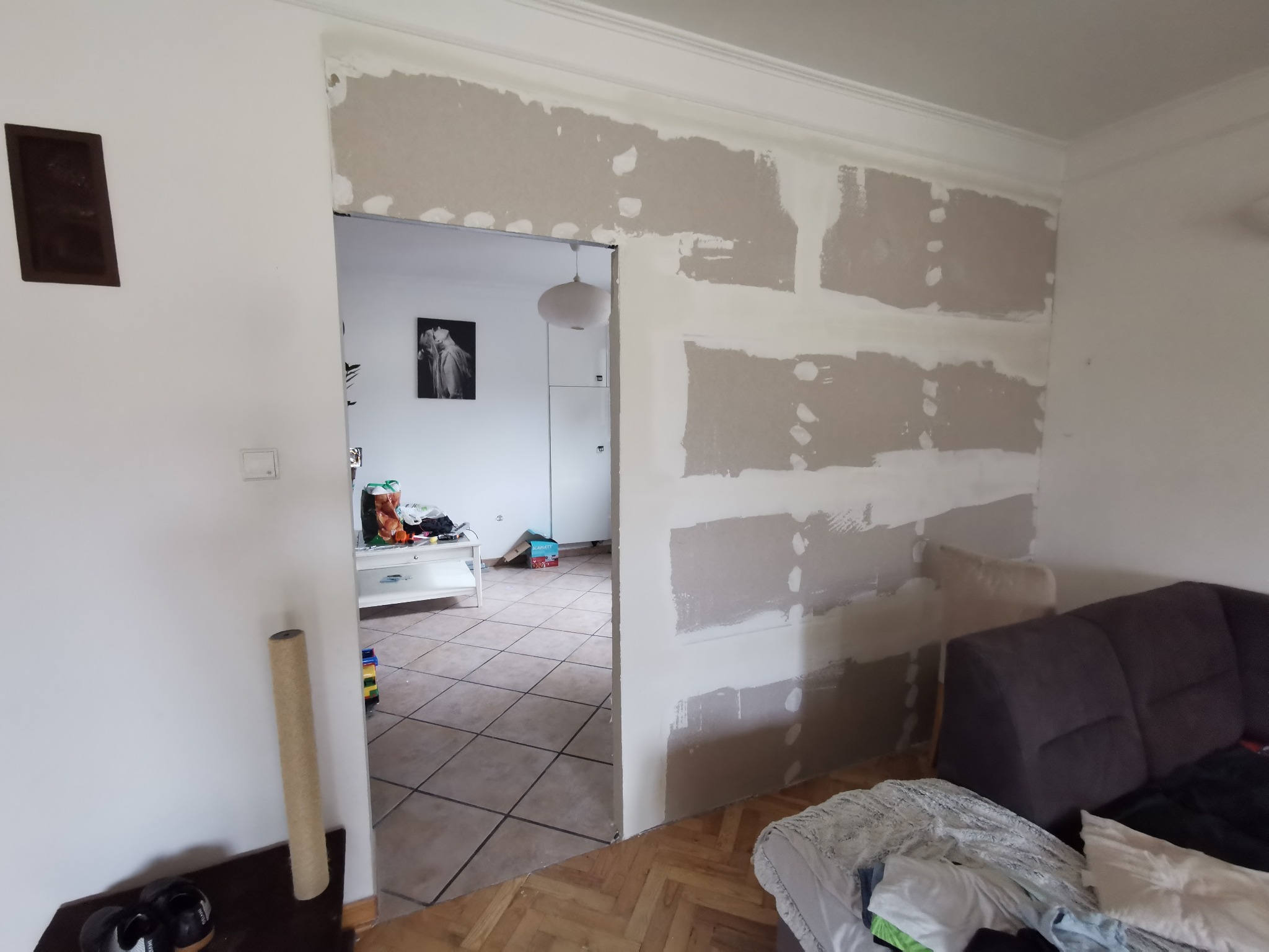 Montaż płyt karton gips na ścianie w trakcie remontu mieszkania, widoczne łączenia płyt wymagające szpachlowania, w tle fragment pokoju z meblami i dekoracjami.