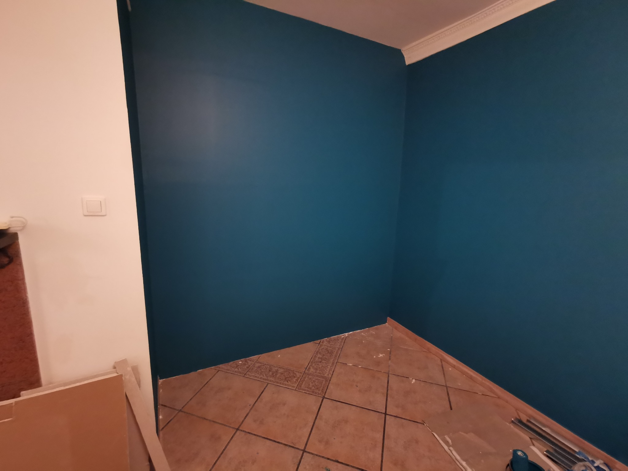 Pomieszczenie po malowaniu ścian na kolor morski, widoczna podłoga z brązowych płytek i biała ściana z włącznikiem światła, na podłodze leżą narzędzia i materiały budowlane.