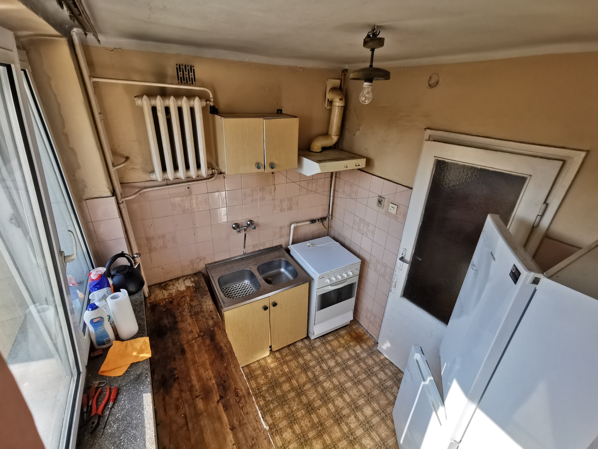 Zdjęcie przedstawia wnętrze zaniedbanej kuchni przed remontem, z widoczną starą zabudową, brudną podłogą, zniszczonymi ścianami i wyposażeniem, w tym kuchenką, zlewem i lodówką.